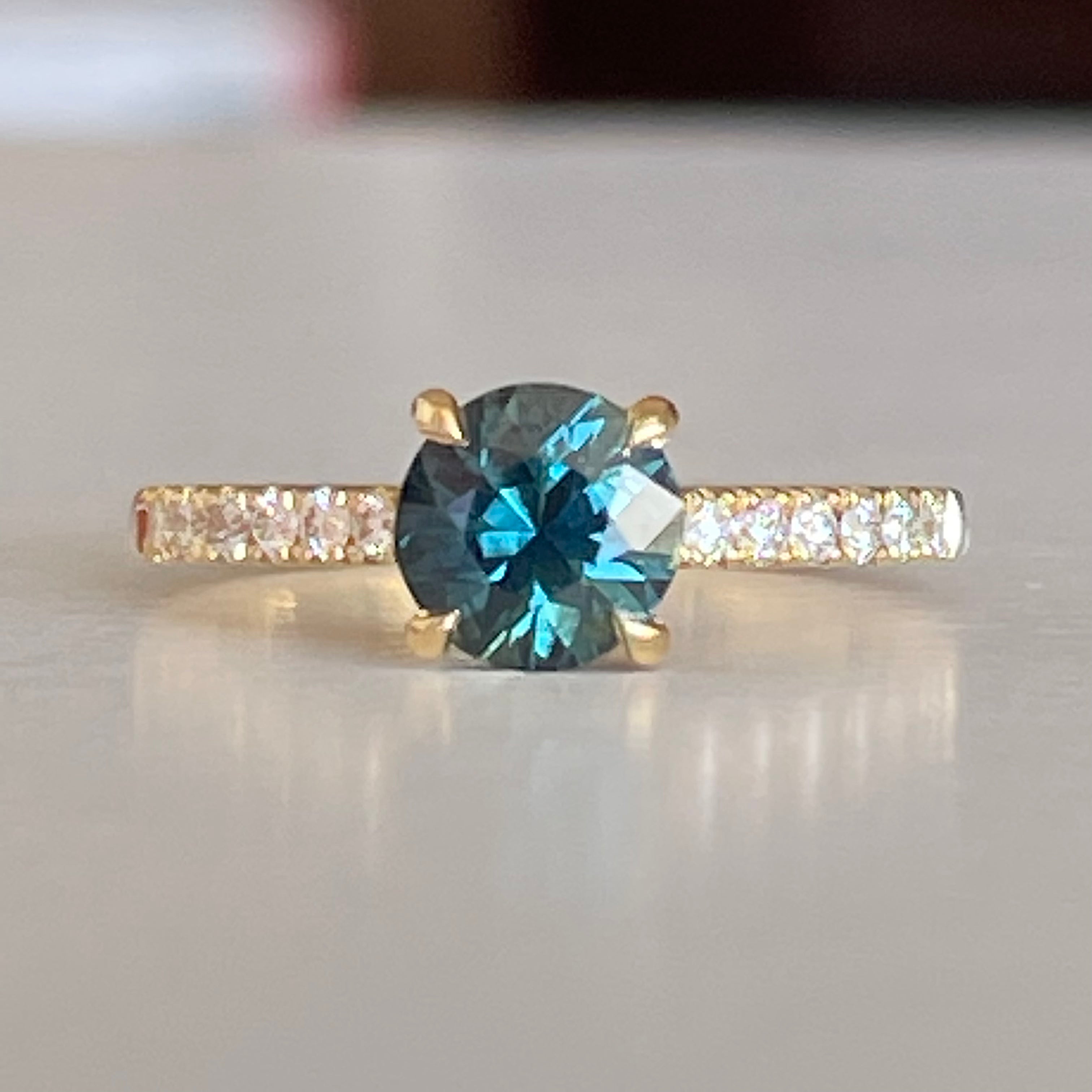 A Blue/Green Australian Parti Sapphire from Rubyvale in QLD & is set with four claws. The stone is beautifully complimented by 12 x Round Brilliant cut total diamond weight 0.18pts and is set in 18ct Yellow gold.  The Sapphire measures 6.4mm and is 1.16cts   Available in Size M  Made with recycled metal in Sydney   Ethically sourced Gemstones